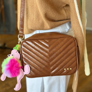 Brown quilted crossbody bag with chevron stitching, gold stud details, and a colorful pink poodle keychain accessory, styled with a tan sweater and white pants.