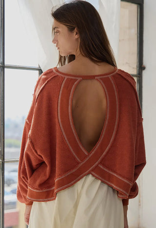 Woman wearing a rust long sleeve top with open back detail and exposed seam standing by a window