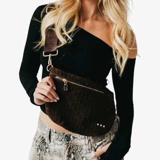 Woman holding a Pretty Simple Suede Westlyn Woven Bum Bag in rich chocolate brown with gold zipper detail, styled with a black one-shoulder top and printed pants.
