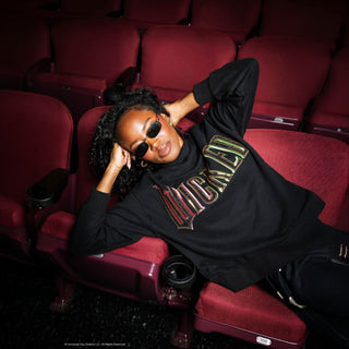 Model reclining in theater seat wearing Queen of Sparkles Licensed Black "Wicked" Logo Sweatshirt