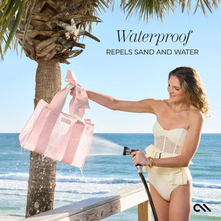 Woman holding a beach bag standing on a beach spraying water on the pink bag with a bow.