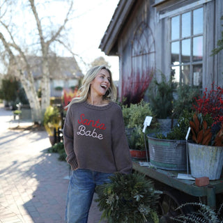 Woman wearing an oversized brown Santa Babe sweater with relaxed denim jeans, holding a holiday wreath while standing outdoors near greenery and rustic décor, showcasing cozy new arrivals for winter