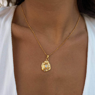 Gold pendant necklace with textured organic charm and crystal center on delicate gold chain worn with white blouse.
