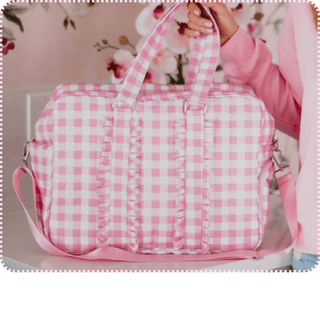 Pink and white gingham duffle bag with ruffle detail, top handles, and adjustable crossbody strap, held in front of a floral background.
