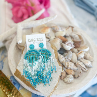 Coastal-inspired beaded fringe earrings in aqua, white, and gold displayed on a stone slab with seashells and starfish in the background.