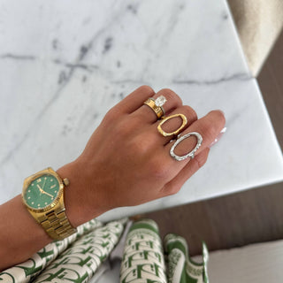 Close-up of a hand wearing a gold open oval ring, paired with a silver open ring and a gold watch with green face, resting on a smooth marble surface.
