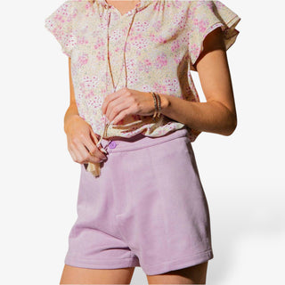 Person wearing a floral top and purple shorts with a button front and pockets on a white background.