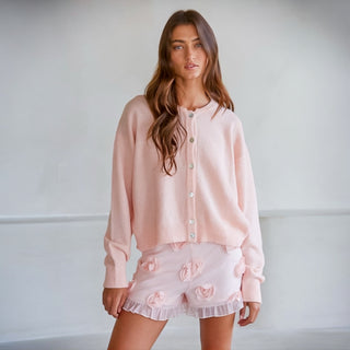 Woman wearing a light pink cardigan and matching shorts with floral details against a plain background