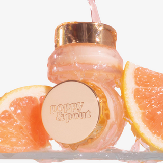 A clear glass bottle with a metallic rose gold cap and matching round lid embossed with the brand name 'poppy & pout', shown on a white background with citrus slices and pink liquid pouring into it.