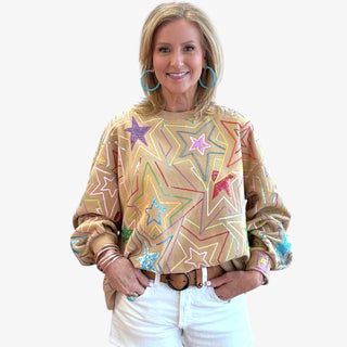 Woman wearing the Queen of Sparkles Multi Star Sweatshirt, a tan oversized pullover featuring colorful sequin star designs in pink, blue, red, purple, and yellow, styled with white shorts and turquoise hoop earrings.