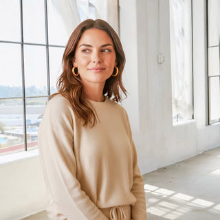 Lady wearing a cream top and pant with gold hoops in a room with white walls and windows