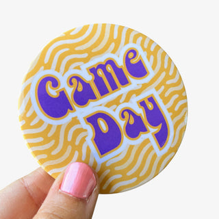 3-inch Game Day button with purple and gold retro lettering on a gold and white tiger stripe background, front view