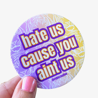 3-inch Hate Us Cause You Ain’t Us game day button with white and gold lettering on a purple and gold tiger-pattern background, front view