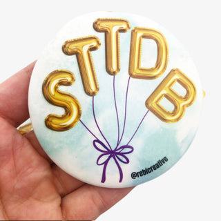 3-inch STTDB game day button featuring gold balloon letters tied with a purple bow on a light blue background, front view