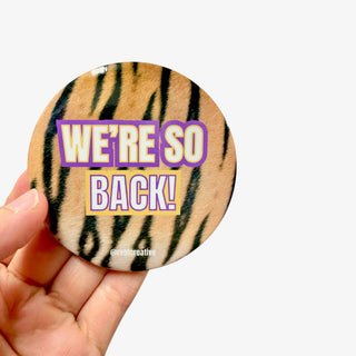 3-inch We’re So Back game day button with purple and gold lettering over a tiger stripe fur background, front view