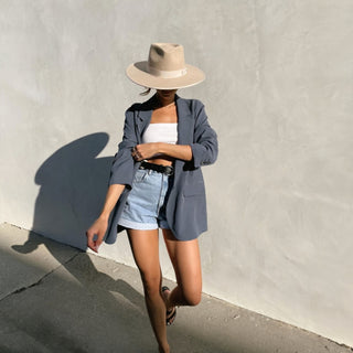 Person wearing a gray blazer, white top, denim shorts, and beige hat against a light gray wall.