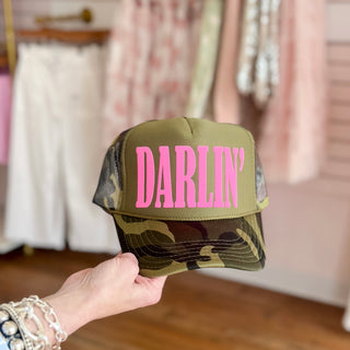 Camouflage cap with 'Darlin' in pink held in front of a clothing store background.