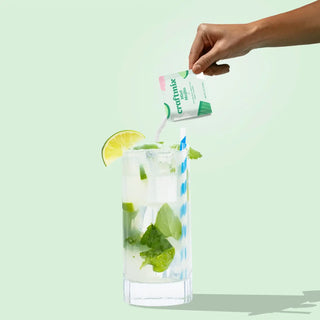 Hand pouring a craftmix mint mojioto packet into a glass of iced drink with lime and mint on a light green background