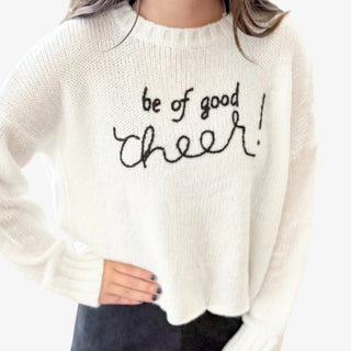 White holiday sweater with black embroidered script reading "Be of Good Cheer!"