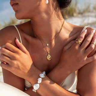 Woman wearing gold jewelry including a necklace, ring, and bracelet with a blurred outdoor background.