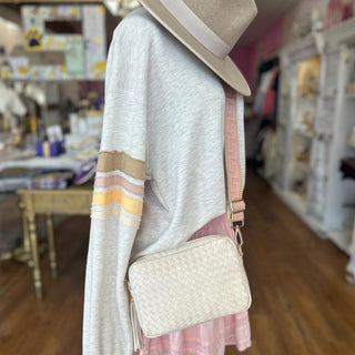 Cream woven crossbody bag with a pink geometric strap and tassel detail, styled with a neutral sweater, pink skirt, and tan wide-brim hat inside a boutique.