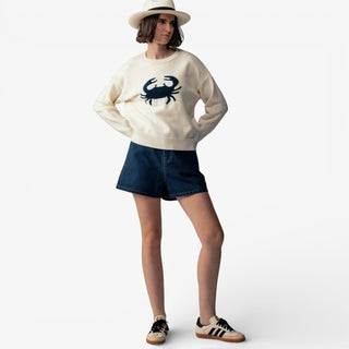 Person wearing a white sweater with a crab design, denim shorts, and a hat on a white background