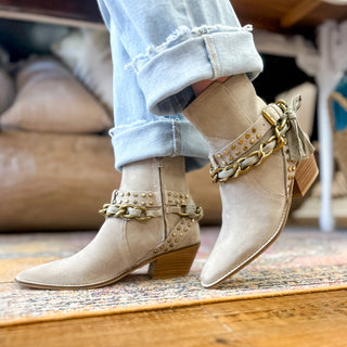 Taupe suede ankle boots with gold stud and chain details, a side tassel accent, and a low stacked heel styled with cuffed light wash jeans for a chic fall look.