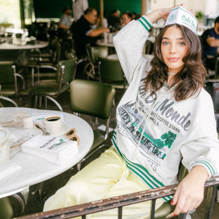 Woman sitting at a cafe table wearing a Queen of Sparkles Cafe Du Monde sweatshirt.