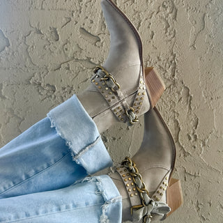 Taupe suede ankle boots with gold studs, chain detailing, and a side bow accent paired with cuffed light wash jeans for a stylish western-inspired look.