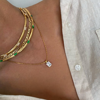 Close-up of a neck with layered gold necklaces: a chunky chain with green stones and a delicate chain with a square white pendant, with a beige shirt.