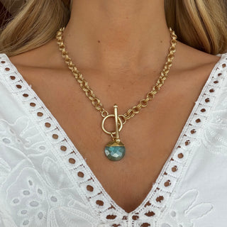 Aquamarine gemstone pendant necklace with chunky gold toggle chain worn on model, faceted blue-green stone pendant close-up front view.