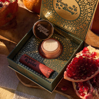Cosmetic set with 'Poppy & Post' branding in an open box, surrounded by pomegranates on a wooden surface.