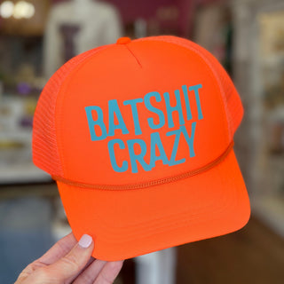 A bright orange trucker hat with turquoise embroidered text reading 'BATSHIT CRAZY' on the front, held by a hand against a blurred indoor background.