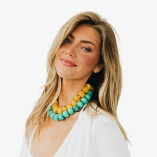 A beaded necklace worn by model in teal green and yellow, made of polished gemstone beads strung on a matching cord, with a small gold clasp. 