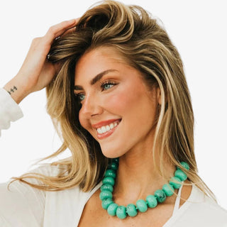 A beaded necklace made of polished dark teal translucent gemstone beads strung on a matching cord, with a small gold clasp, shown against a light gray background.