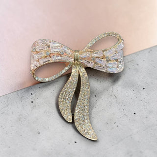 Gold bow-shaped brooch with clear gemstones on a light pink and gray surface