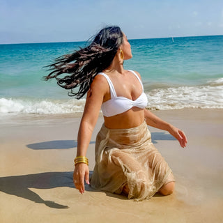BudhaGirl All Weather Gold Bangles stacked on wrist with white swimsuit and beach cover-up, ocean shoreline lifestyle view