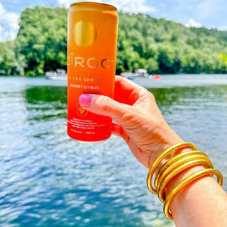 BudhaGirl All Weather Gold Bangles stacked on wrist holding citrus spritz drink by the lake, waterproof luxury bracelet set lifestyle view