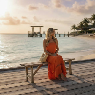Woman wearing orange scallop trim strapless maxi dress sitting on beach boardwalk at sunset with woven handbag