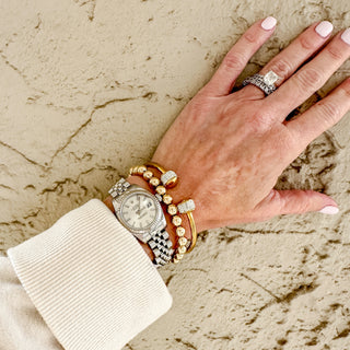 Hand wearing multiple bracelets, a watch and a gold cuff with cz design end caps on a textured beige surface
