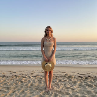 Model wearing coastal gold and pearl charm necklace with strapless linen dress on beach at sunset, full-body lifestyle view