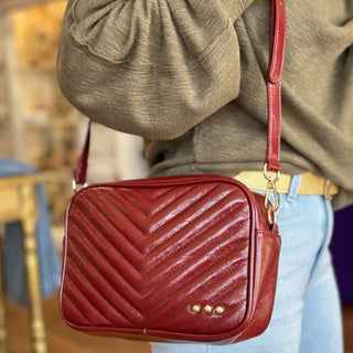 Deep red quilted crossbody bag with chevron stitching, gold stud accents, and adjustable strap, styled with an olive sweater and light wash jeans.