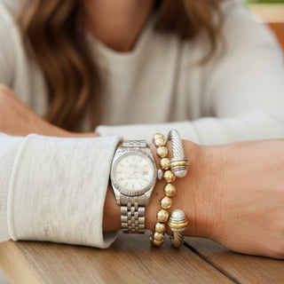 Close-up of a wrist wearing a silver watch and gold bracelets on a wooden surface.
