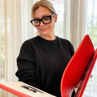 Woman wearing EyeJets Dubai black velvet blue light reading glasses while reading a large red book in a bright, naturally lit room.