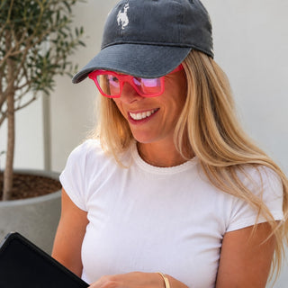 EyeJets Ibiza pink blue light reading glasses worn by a smiling woman with a gray baseball cap and white tee in a bright outdoor lifestyle setting.