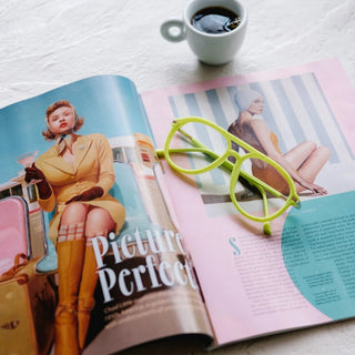 EyeJets Mykonos neon green velvet reading glasses styled on pink magazine with coffee flat lay lifestyle view