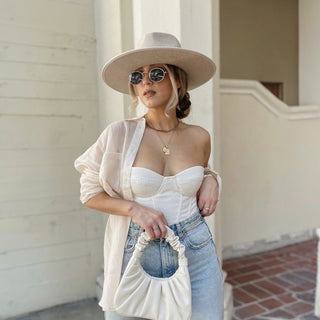Woman wearing a beige hat, sunglasses, and a white outfit with blue jeans, standing against a light-colored wall.