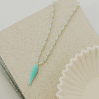 Aqua fish pendant necklace with ivory beaded chain and gold accents displayed on neutral linen background flat lay view.