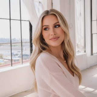 A blonde woman with wavy hair wearing a pale pink knit sweater, posing in a bright, modern room with large windows.