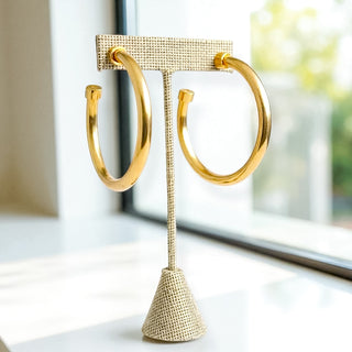 Large gold hoop earrings with flat stud ends, displayed on a textured beige earring stand near a sunlit window